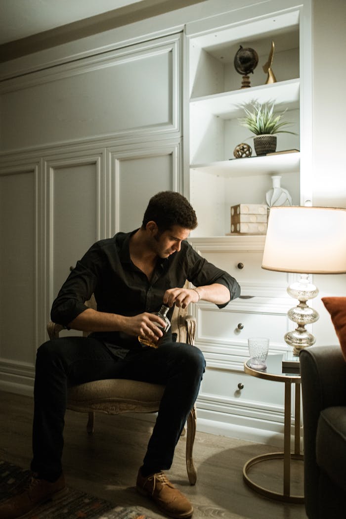 A man sits contemplatively with a drink in a warmly lit living room setting.